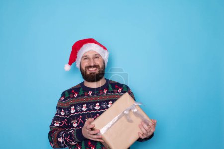 Photo for Attractive bearded man in Santa hat with gift box on blue background. Christmas or New year concept. - Royalty Free Image
