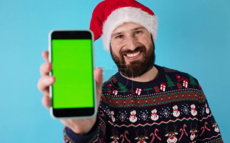 Photo for Merry bearded man wear knitted christmas sweater posing holding mobile cell phone with blank screen workspace area isolated on blue background. Happy New Year 2024 holiday concept. - Royalty Free Image