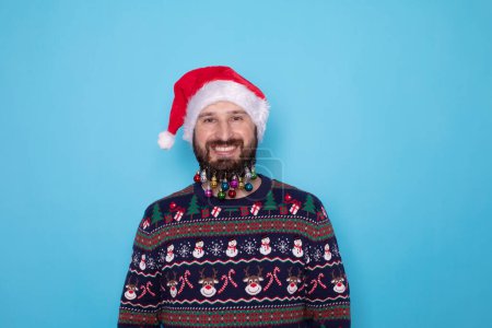 Photo for Happy young bearded man in Christmas sweater and Santa hat smiling at camera on light blue background. - Royalty Free Image