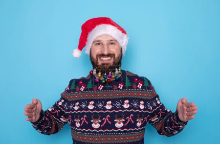 Photo for Happy young bearded man in Christmas sweater and Santa hat showing something on light blue background. Space for text. - Royalty Free Image