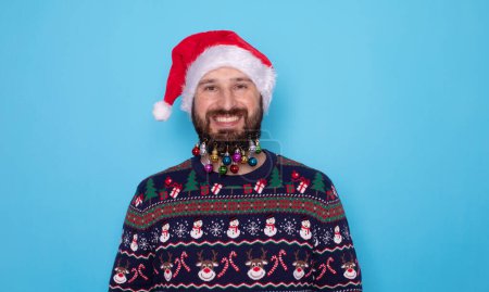 Téléchargez les photos : Joyeux jeune homme barbu en pull de Noël et chapeau de Père Noël souriant à la caméra sur fond bleu clair. - en image libre de droit