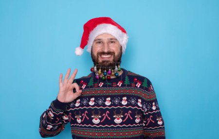 Téléchargez les photos : Heureux jeune homme barbu en pull de Noël et chapeau de Père Noël montrant ok signe sur fond bleu clair. - en image libre de droit
