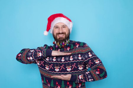 Photo for Happy young bearded man in Christmas sweater and Santa hat showing something on light blue background. Space for text. - Royalty Free Image