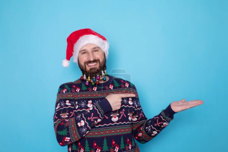 Foto de Feliz joven barbudo en jersey de Navidad y sombrero de Santa señalando algo sobre fondo azul claro. Espacio para texto. - Imagen libre de derechos