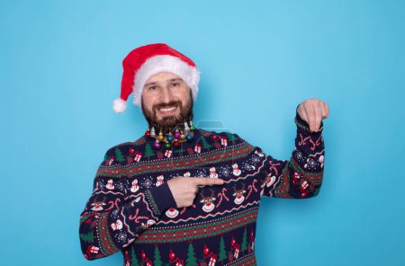 Photo for Happy young bearded man in Christmas sweater and Santa hat pointing at something on light blue background. Space for text. - Royalty Free Image