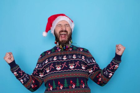 Foto de Retrato de hombre barbudo atractivo emocional celebrando año nuevo, vistiendo suéter y sombrero de santa, gritando de alegría, ganando o triunfando, de pie sobre fondo azul. - Imagen libre de derechos