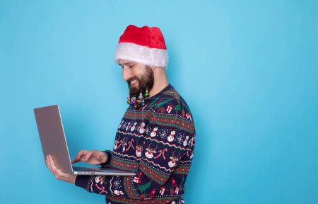 Téléchargez les photos : Homme IT barbu portant un pull tricoté et un chapeau de Père Noël posant tenant à l'aide d'un ordinateur portable isolé sur fond bleu. - en image libre de droit