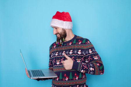 Photo for Bearded IT man wearing knitted sweater and Santa hat posing holding using laptop pc computer isolated on blue background. - Royalty Free Image
