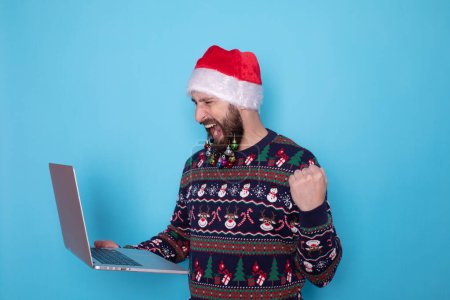 Téléchargez les photos : Homme IT barbu portant un pull tricoté et un chapeau de Père Noël posant tenant à l'aide d'un ordinateur portable isolé sur fond bleu. - en image libre de droit