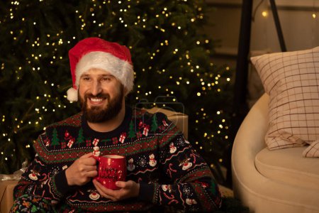 Téléchargez les photos : Jeune homme barbu buvant du café près de l'arbre de Noël à la maison. Noël à la maison. - en image libre de droit