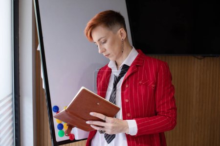 Foto de Retrato de una hermosa mujer de negocios inteligente de pelo rojo o gerente, de pie en la oficina moderna, vestido con ropa elegante, sosteniendo el cuaderno en las manos. - Imagen libre de derechos
