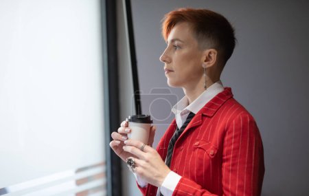 Téléchargez les photos : Portrait d'une belle femme d'affaires ou d'un manager aux cheveux roux confiant, debout dans un bureau moderne, vêtu de vêtements élégants, tenant une tasse de café dans les mains. - en image libre de droit