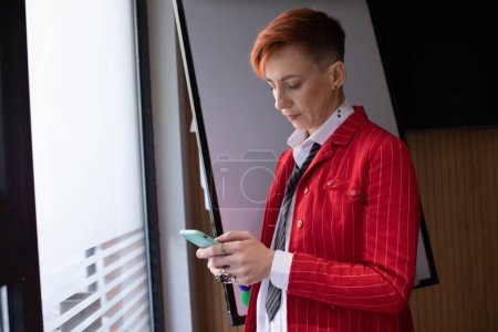Foto de Retrato de una hermosa mujer de negocios inteligente de pelo rojo o gerente, de pie en la oficina moderna, vestida con ropa elegante, usando un teléfono inteligente - Imagen libre de derechos