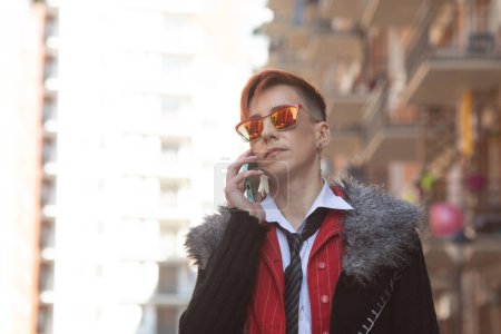 Foto de Hermosa mujer de mediana edad en traje rojo y gafas de sol hablando en el teléfono inteligente contra la calle. - Imagen libre de derechos