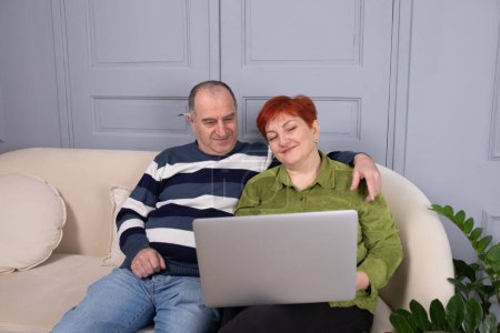Téléchargez les photos : Couple d'âge moyen assis sur le canapé et travaillant à la maison sur ordinateur portable. - en image libre de droit