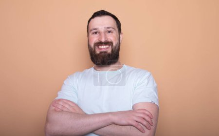 Photo for Handsome bearded young man posing with crossed arms and smiling at camera isolated on beige background - Royalty Free Image