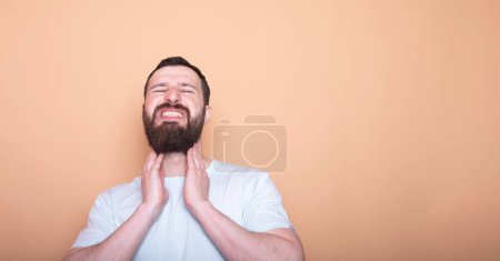 Photo for Young bearded man having sore throat and touching his neck, wearing white t-shirt against light beige background. - Royalty Free Image