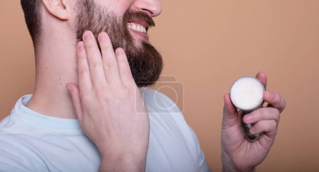 Hombre barbudo guapo presentando bálsamo de barba sobre fondo beige