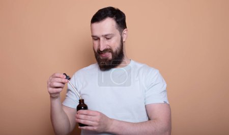 Photo for Handsome bearded man presenting beard oil on beige background - Royalty Free Image