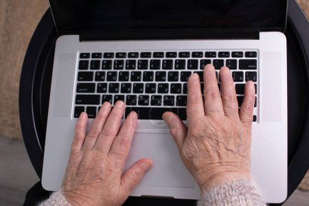 Téléchargez les photos : Vue rapprochée d'une femme âgée utilisant un ordinateur au bureau à domicile. Concept de télétravail. Femme âgée acheter en ligne, passer du temps libre sur Internet, profiter de la communication dans les médias sociaux, se reposer à la maison. - en image libre de droit