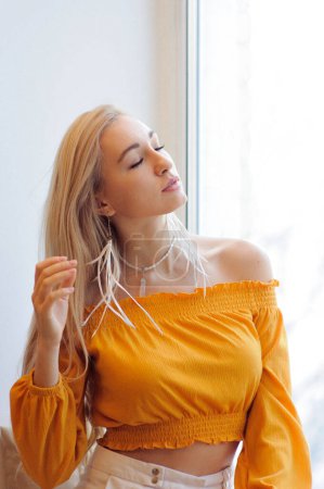 Photo for Portrait of gorgeous blonde woman in boho chic yellow blouse and long white feather earrings sitting near window - Royalty Free Image