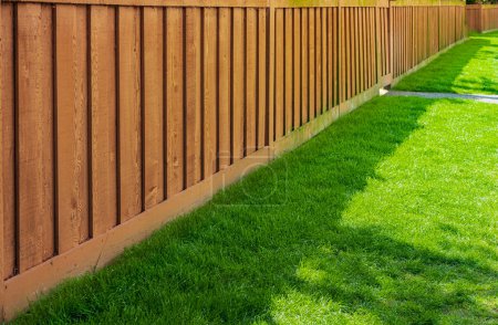 Nice new wooden fence around house. Wooden brown fence with green grass lawn. Street photo, nobody, selective focus