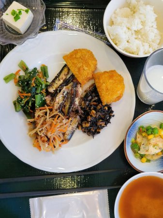 Authentic Japanese breakfast, featuring a varied plate with sardines, croquettes, vegetables, and legumes, accompanied by miso soup, tofu, Japanese rice (gohan), and traditional chopsticks (hashi). Perfect image to illustrate articles about Japanese 