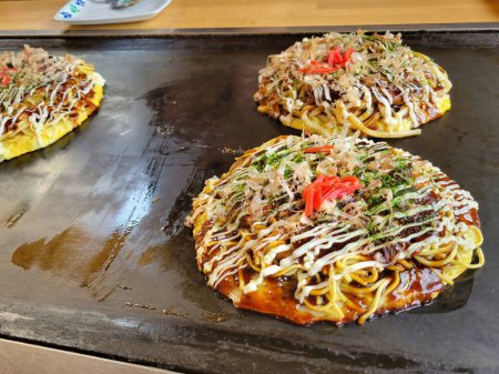 Un chef prépare savamment un Okonomiyaki salé, un plat japonais populaire d'Okayama, sur une plaque de cuisson en acier chaud, remplissant l'air d'un arôme appétissant, dans un restaurant japonais traditionnel au Japon.