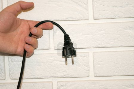 Foto de Vista de una mano sosteniendo un enchufe negro 220v contra una pared de ladrillo blanco - Imagen libre de derechos