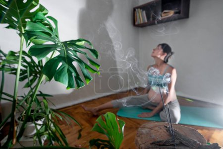 Foto de Pregnant woman sits on a yoga mat in a meditation pose. Pregnant woman doing yoga exercises at home. Incense room for relaxation during yoga - Imagen libre de derechos