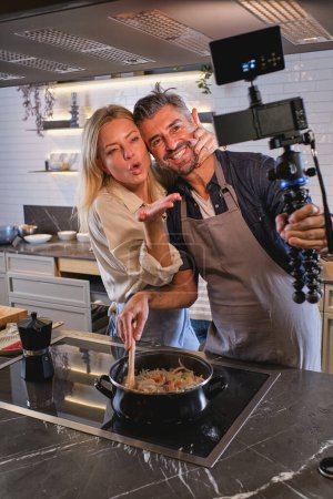 Téléchargez les photos : Aimer couple joyeux debout ensemble près de la table de cuisson et de prendre selfie tout en cuisinant le dîner traditionnel dans la cuisine moderne - en image libre de droit