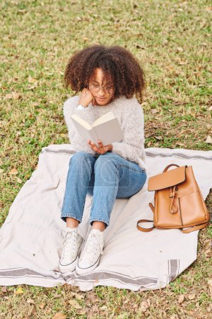 Foto de Longitud completa de la mujer negra enfocada en ropa casual sentada en manta y apoyada en la mano mientras lee una novela interesante en la naturaleza - Imagen libre de derechos