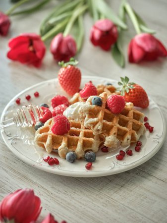 Photo for From above of delicious waffles with ripe berries and honey dipper served on table with red tulips in light kitchen - Royalty Free Image