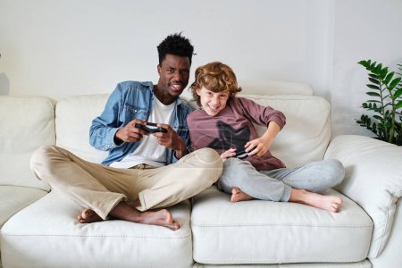 Téléchargez les photos : Corps plein joyeux jeune noir mâle et mignon adolescent garçon jouer jeu vidéo ensemble tandis que assis avec les jambes croisées sur canapé avec joysticks - en image libre de droit