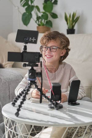 Téléchargez les photos : Enfant ravi en cours d'enregistrement vidéo pour blog en ligne tout en étant assis à la table blanche avec caméra sur trépied et microphone à la maison - en image libre de droit