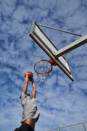 Photo for From below side view of sportive guy in activewear doing slam dunk on outdoor sports ground during basketball workout - Royalty Free Image