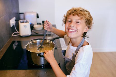 Téléchargez les photos : Vue latérale de l'enfant joyeux dans le tablier regardant la caméra tout en remuant les pâtes dans une casserole sur le poêle à la maison - en image libre de droit