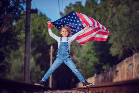 Téléchargez les photos : Corps complet de garçon blond levant le drapeau américain flottant dans les mains et regardant fièrement la caméra tout en se tenant debout sur le chemin de fer - en image libre de droit