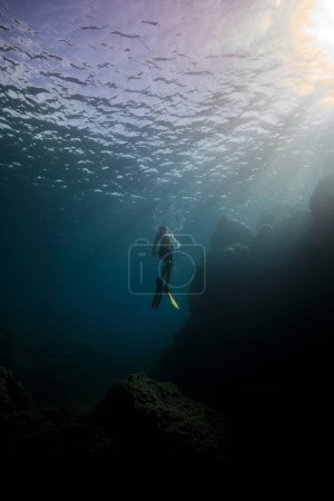 Téléchargez les photos : Corps complet de plongeur mâle méconnaissable avec palmes et masque de plongée nageant dans l'eau de mer profonde sombre près du fond pierreux et regardant vers le haut sur le soleil - en image libre de droit
