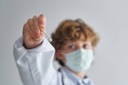 Photo for Kid in medical uniform and disposable mask showing cotton swab on white background in daytime - Royalty Free Image