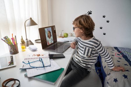 Téléchargez les photos : Vue latérale du garçon mignon dans les lunettes vidéo bavardage avec un camarade de classe sur ordinateur portable à la table avec des cahiers d'exercices dans la salle de lumière - en image libre de droit