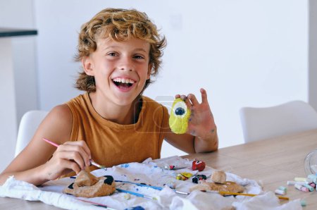 Téléchargez les photos : Enfant préadolescent positif montrant un caillou peint vibrant assis à la table et regardant la caméra - en image libre de droit