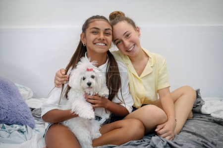Foto de Sonriendo diversas amigas adolescentes sentadas en la cama y abrazando al perro mientras miran la cámara - Imagen libre de derechos