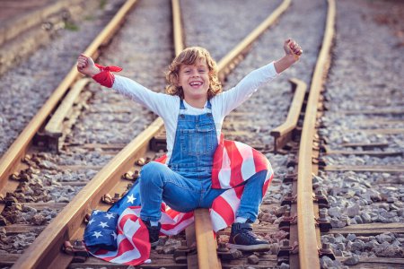 Téléchargez les photos : Corps complet de garçon souriant regardant la caméra avec les bras levés tout en étant assis sur le chemin de fer en métal avec le drapeau coloré des États-Unis - en image libre de droit