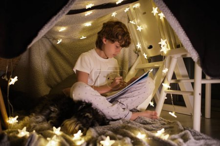 Photo for Full body of talented boy drawing in sketchbook while sitting with crossed legs near dog in cozy room with glowing garland - Royalty Free Image