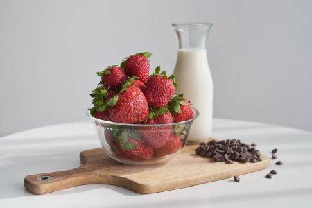 Photo for Delicious ripe strawberries and chocolate chips with glass bottle of milk placed on wooden chopping board on white round table - Royalty Free Image