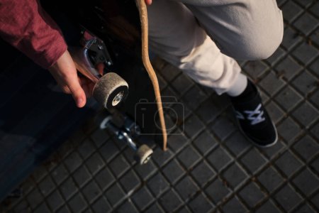 Téléchargez les photos : D'en haut garçon anonyme avec planche à roulettes debout sur le trottoir pavé et reposant sur la rue de la ville - en image libre de droit