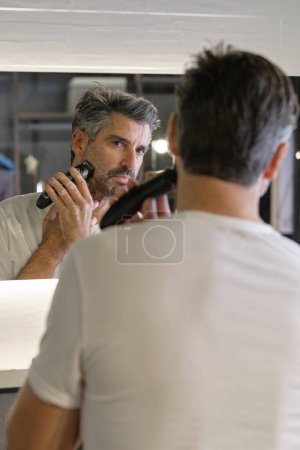 Photo for Focused man in white t shirt looking at reflection in mirror and shaving beard with trimmer in modern bathroom - Royalty Free Image