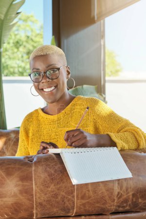 Téléchargez les photos : Portrait de femme afro-américaine souriante écrivant dans un journal assis sur un canapé à la maison - en image libre de droit