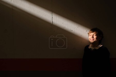 Téléchargez les photos : Positif mignon preteen garçon debout avec les yeux fermés au mur dans chambre sombre attrapant rayon de soleil dans le visage - en image libre de droit